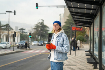 Long-haired hipster guy in stylish outfit, standing in bus stop with cup of coffee, looking into distance, waiting for taxi, bus. Carefree fashionable male, representative of generation z - obrazy, fototapety, plakaty
