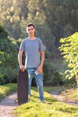 Portrait of a skater man smiling and looking at camera. He is holding his longboard next to him. It is in an area of trees and natural landscapes - obrazy, fototapety, plakaty