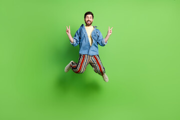 Full length photo of handsome cheerful guy wear denim jacket jumping high showing v-signs isolated green color background - obrazy, fototapety, plakaty