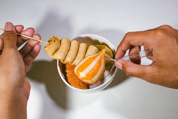 a porcelain bowl containing odeng and fishcake, where the odeng is held in the left hand and the...