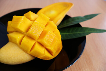 A ripe mango cut in half sits on top of another fruit and leaves in a black porcelain plate at a wooden table.

