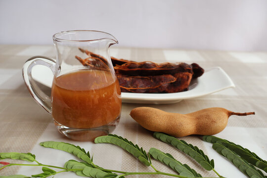 Glass jar with concentrated tamarind juice. Ripe fruit is placed on the side and peeled on a plate next to a white background.

