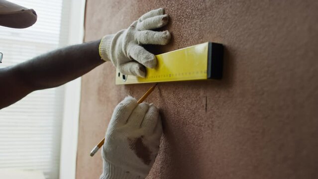 Side closeup of Black male contractor making marks on brown wall with pencil using waterpas measurement tool before hanging shelf or other furniture in remodeled room