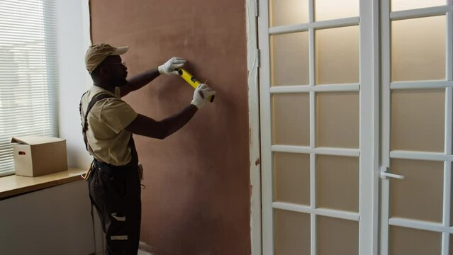 Side full medium footage of Black male contractor in cap and workwear making marks on wall using measurement tool during room renovation