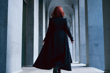 Young woman wearing long black cloak with red hair walking with her back turned in corridor between...
