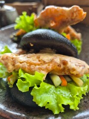 A plate of food with a black bun and lettuce. The bun is topped with a sauce and a piece of salted egg soft shell crab. Selective focus.