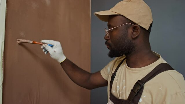 Side tilt footage of serious diligent Black repairman in cap and workwear painting wall brown using paintbrush and holding tray in hands while working in house