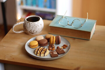 Plate with macarons, cookies, chocolate and nuts, cup of tea or coffee, book and reading glasses on the table. Selective focus. Colorful bookcase in the background.
