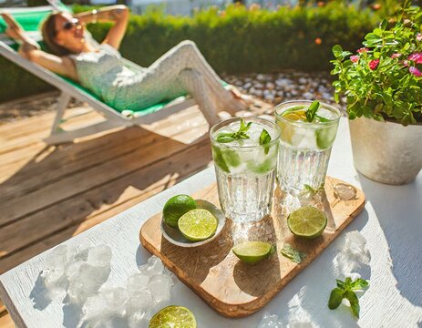 Bezalkoholowe mojito. Szklanki z orzeźwiającym napojem z limonką, miętą i kostkami lodu stojące na stole na tarasie. W tle widać siedzącą na leżaku kobietę kt&oacute;ra się opala. Wakacyjny relaks