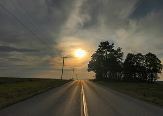 Looking into the setting sun on a two-lane black top road with power lines and pine trees silhouetted