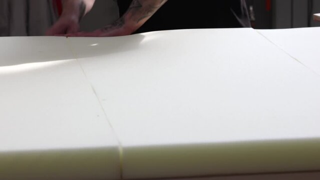 A worker in a furniture factory lays thick sheets of foam rubber tightly together. Male hands with tattoo close-up tamping foam sheets in furniture factory