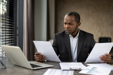 Fototapeta premium Sceptic formal male in business suit looking attentively on sheets of paper in both hands while sitting by desktop. Focused accountant comparing data on two documents from different departments.