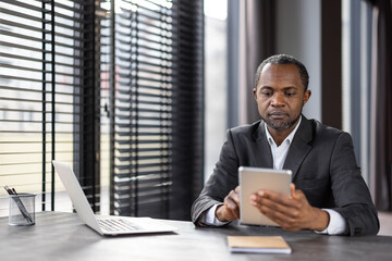 Focused entrepreneur scrolling internet through digital tablet while working by window with jalousie. Attentive financial manager entering data about company bills in corporate system application.