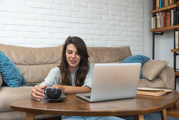 Young freelance business woman graphic designer taking a break from working at home and talk on video call with friend while drinking tea or coffee and enjoying in conversation.