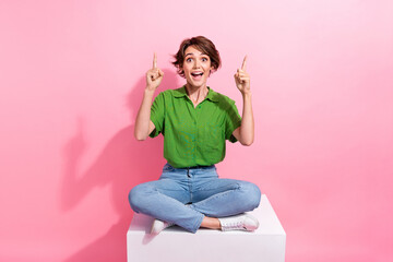 Naklejka premium Full body photo of astonished pretty lady sit podium indicate fingers up empty space offer isolated on pink color background