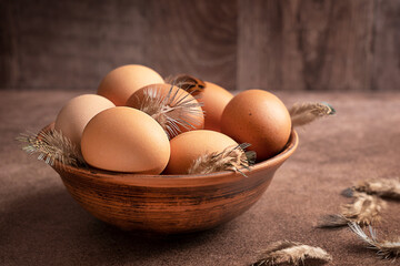 Heap of raw uncooked whole chicken eggs with hen feathers in ceramic brown bowl on table against wooden wall freshly gathered in henhouse as healthy organic ingredient in culinary full of protein