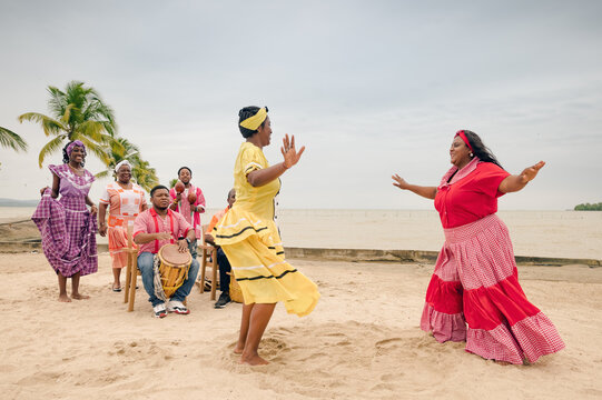 Two young women dressed in vibrantly colored clothing dance to the beat of drums at a beach party.