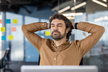 Calm caucasian guy sitting on chair with hands behind head and closed eyes in background of stylish office. Digital designer daydreaming and setting mind up for creative mood at beginning of workday.