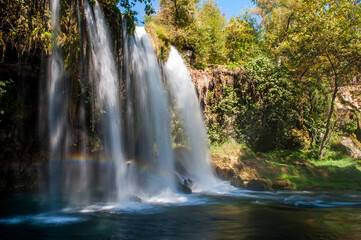Duden Waterfall cascading into a blue pool with a visible rainbow, surrounded by rich green foliage in Antalya, Turkey.