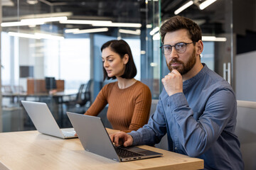 Pensive unshaved guy in denim shirt resting head on hand while looking at camera next to female colleague. Two digital designers developing new creative strategy for social media for marketing team.