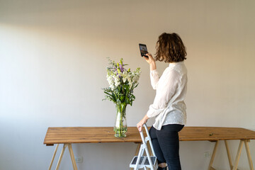Woman taking photos flower arrangement on wooden table.