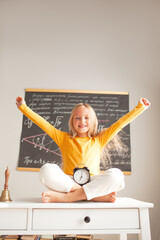 a little girl 6-7 years old is getting ready for school, the girl is dressed in a yellow long sleeve and sitting at a desk against the backdrop of desk, the kid holding alarm