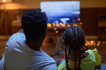 Back view of Black family father and daughter watching Tv together in dark room sitting on couch