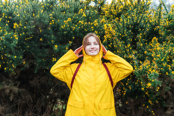 Smiling young woman in a yellow raincoat and hood enjoying walk on background of blooming bush with flowers. Travel concept. Hiking in any weather. Girl on a journey. Relaxing, personal fulfillment