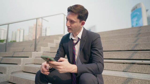 Young salary office man sit down at the stairs looking at cellphone showing disappointed face after reading bad news on the phone screen, thing goes wrong not going as planned, got fired from company