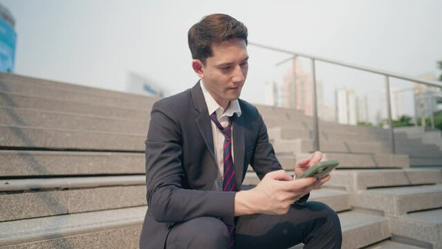 Young salary office man sit down at the stairs looking at cellphone showing disappointed face after reading bad news on the phone screen, thing goes wrong not going as planned, got fired from company,
