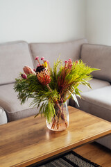 Vase with exotic protea flowers bouquet on coffee table with blurred background of modern cozy light living room with gray couch sofa. Open space home interior design. Elegant home decor. Copy space.