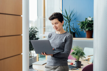Smiling Neutral gender middle aged woman in casual outfit working on laptop while standing in modern office, meeting room, conference space. Business process organization. Comfy workplace.