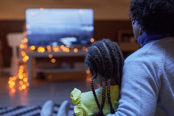 Back view of Black mother and daughter watching Tv together in dark room sitting on couch and embracing copy space