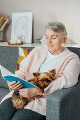 Elderly retired senior woman with wrinkles smiling while embracing her Yorkshire terrier dog pet and relaxing while reading on sofa at home. Best friend. Enjoying retirement lifestyle. Vertical card.