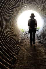 Woman from behind with hat, backpack and hiking sticks going through a dark tunnel with the light...