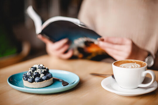 A woman relishes a tranquil moment, engrossed in a book while savoring a blueberry tart and coffee. The scene exudes relaxation and leisure, a delightful blend of literature and culinary indulgence.