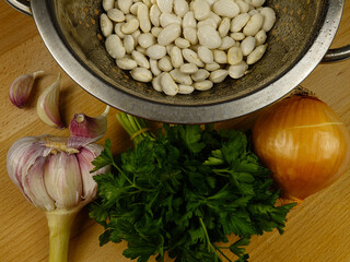 Soaked white beans in a colander, a bunch of parsley, an onion and garlic lie on a wooden table. Ingredients for preparing a bean dish.
