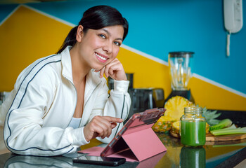 Healthy Morning Routine: Confident Latina Woman Balances Fitness and Work in Her Bright Kitchen, Enjoying Freshly Cut Fruit and Green Juice After a Workout. 