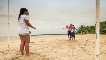 A girl watches two boys fight for the ball in a soccer game on a beach.
