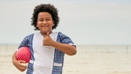 Portrait of boy on the beach holding a ball, looking at camera with his thumb up.