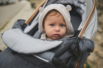 Portrait of an adorable baby boy with hood on his head, looking at the camera, sitting in a baby stroller outdoors