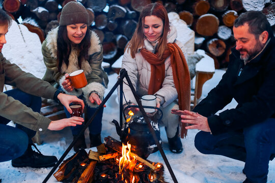 Group of friends gathering around bonfire in backyard, drinking tea and warming hand over fire