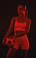 A beautiful athletic blonde girl in white shorts and a top plays football on a black background in red light.