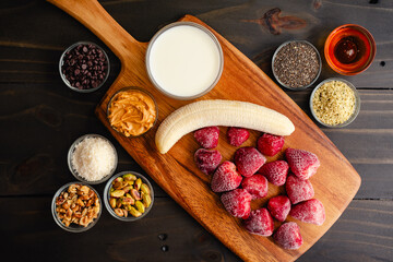 Strawberry Smoothie Bowl Ingredients on a Wooden Cutting Board: Frozen strawberries and banana with milk and various smoothy bowl toppings