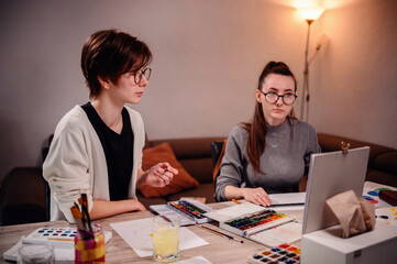 Two artists deeply focused, one paints while the other evaluates, amidst a home office setting...