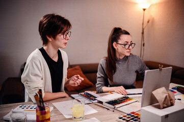 Two artists deeply focused, one paints while the other evaluates, amidst a home office setting...