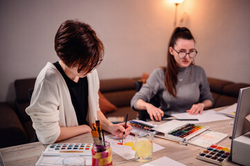 Fototapeta premium Two concentrated women involved in an artistic project, with one painting and the other measuring, surrounded by creative tools