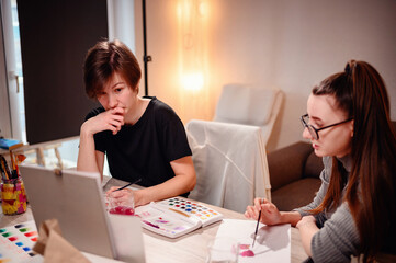 Two artists engage in a collaborative discussion over a watercolor palette, with one offering...