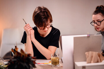 Artists share a moment of collaboration, one meticulously mixing paint while the other evaluates...
