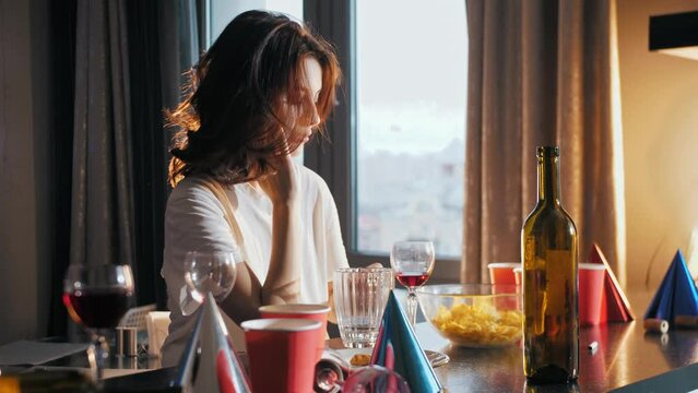 Hangover. Young girl, after a night of partying, is sitting at a table amidst the chaos and mess of her surroundings, nursing a hangover. With a pounding headache, she tries to gather her thoughts and
